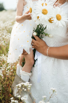 ender moment of a mother caring for her cute little daughter, adjusting her white headscarf and styling small flowers in her hair while sitting together in a wild meadow filled with white flowers. 