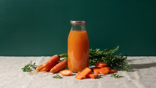 A glass bottle filled with fresh organic carrot juice surrounded by raw whole and sliced carrots on a rustic surface