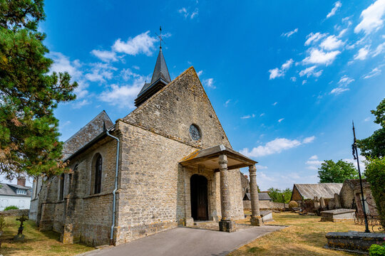 Historic Normandy stone church with a pointed slate spire and cross, photographed from a low angle against a bright blue sky with scattered clouds, a pine tree and small graveyard visible in the foreg