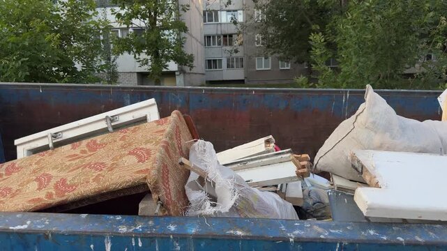 Blue dumpster filled with old mattress, wooden frames and white sacks near an apartment building. waste, disposal, urban, cleanup, renovation