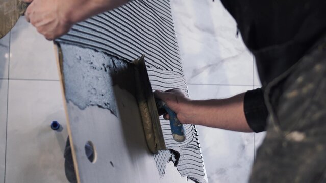 Master tiler spreads glue on huge large format tiles of XXL size. Notched trowel, professional tile work, bathroom renovation, laying blue tiles, slow motion