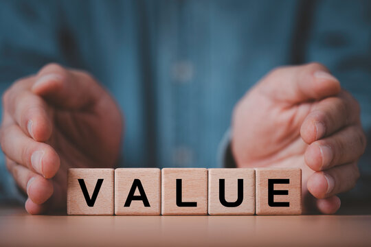 Hands protecting wooden blocks spelling VALUE on a desk, symbolizing value protection, brand value, business security, investment, customer trust, asset management, and long-term business success.