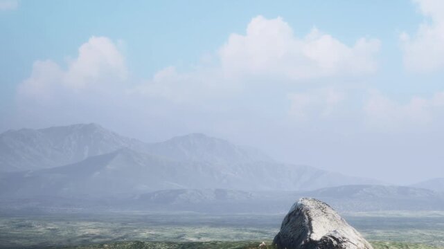 A large rock sits in the foreground while mountains rise in the background under a clear sky. Clouds linger above, creating a vast landscape scene during daylight.