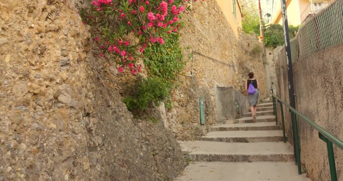 Female tourist with purple backpack climbs steep stone steps between rustic village walls in Roquebrune Cap Martin.