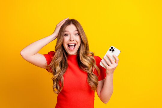 Young woman in red dress excitedly reacts while holding a smartphone against a bright yellow background, surprised joyful expression and modern mobile lifestyle moment