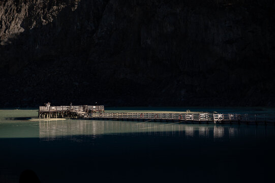 Horizontal view of illuminated wooden pontoon pier on turquoise sulfur lake under dark volcanic cliff at Kawah Putih crater, Ciwidey, West Java, Indonesia.