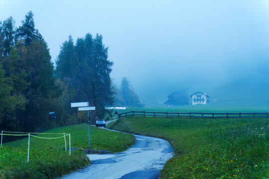 Foggy Rural Road Winding Through Green Meadows Past Alpine Farmhouse at Morning