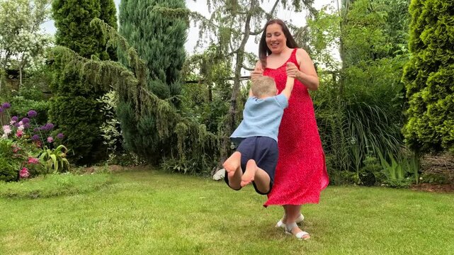 Mother spins a laughing boy around in a summer garden, joyful family play, active childhood, parent and child bonding, outdoor lifestyle.
