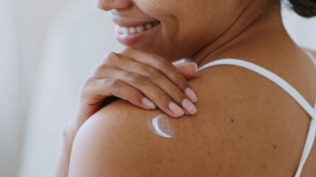 Smiling Young Black Woman Applying Moisturizer To Her Skin With Hand While Looking Back At Camera, Relaxing In Bathroom At Home. Home Skincare Concept.