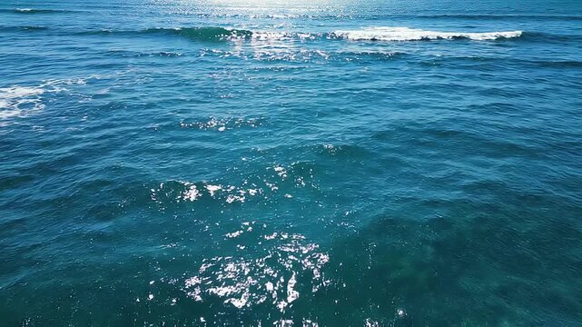 Vista a&eacute;rea del mar con intensas olas azules rompiendo en espuma blanca y destellos de luz solar sobre el agua.