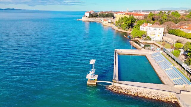 Drone flying backwards from Kolovare&rsquo;s jump platform and pool in spring, revealing the calm Adriatic and distant Zadar old town in soft early‑season light.