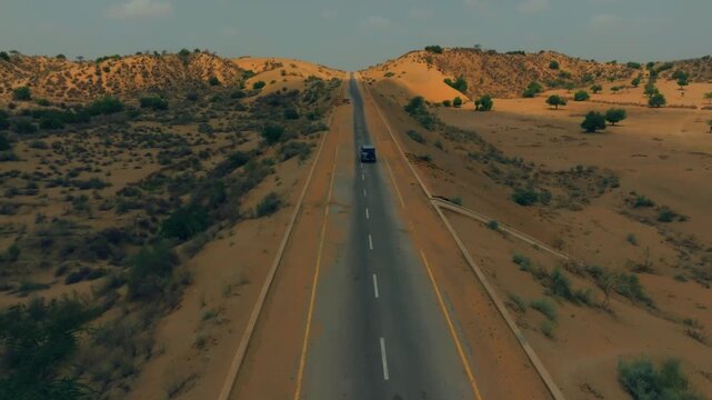 Expansive aerial shot showcasing a car traveling on a long road cutting through the arid and majestic Thar Desert landscape of Sindh, Pakistan.