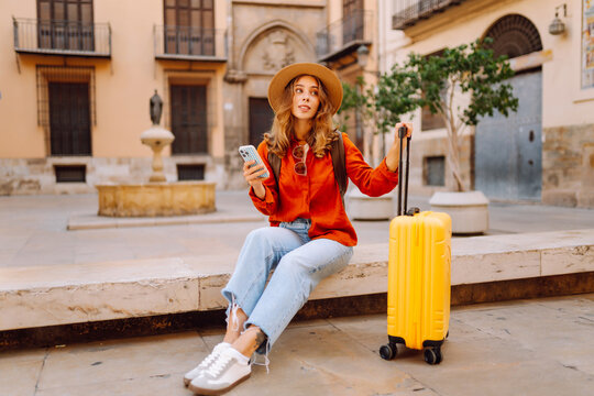 Tourist wearing hat and carrying a brightly colored suitcase sits in town square, holding phone. A young woman with a smartphone is relaxing outdoors. Concept for tourism, weekends, or vacations.
