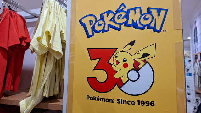 yellow promotional sign featuring pokemon pikachu 30th anniversary logo next to colorful t shirts hanging on rack inside Uniqlo retail store on July 21, 2026 in Bangkok, Thailand.