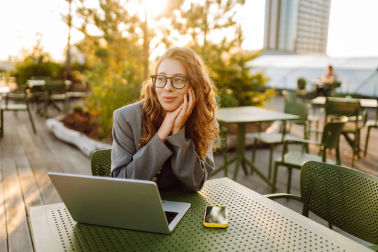 Young woman sits at table with a laptop on summer terrace. Stylish woman works on laptop, types, and enjoys sunset outdoors. Concepts for remote work, freelancing, and relaxation.