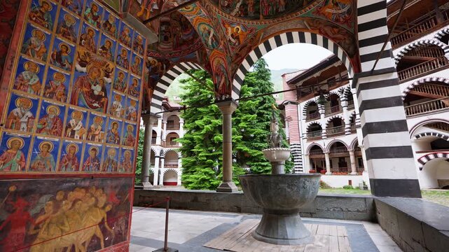 Rila Monastery courtyard with Orthodox frescoes, religious icons, stone fountain and striped arches in Bulgaria, historic Christian architecture during summer travel