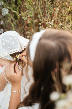 ender moment of a mother caring for her cute little daughter, adjusting her white headscarf and styling small flowers in her hair while sitting together in a wild meadow filled with white flowers. 