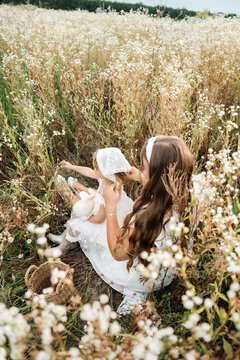 ender moment of a mother caring for her cute little daughter, adjusting her white headscarf and styling small flowers in her hair while sitting together in a wild meadow filled with white flowers. 