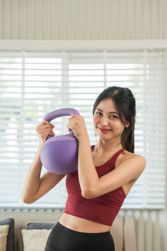 Attractive asian young fitness woman lifting kettlebell dumbbell weights workout at home in living room. Fresh feeling female training and exercise with kettlebell wearing sport wear fit body.