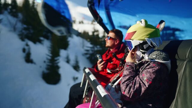 A father and child are riding a ski lift up a snowy mountain. The pair ascend the slopes, enjoying the winter landscape.