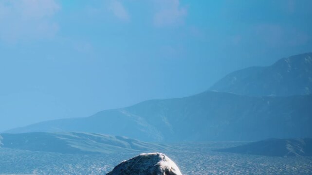 A person stands on a rock while hiking in a mountainous area. The clear sky shows no clouds and hills are visible in the background. Sunlight brightens the scene.