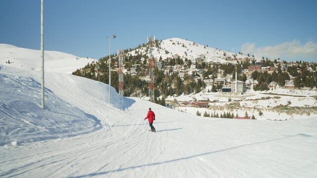 A lone snowboarder in a red jacket rides down a wide, pristine mountain slope. Enjoying a thrilling winter sport experience on a clear, sunny day at a ski resort.