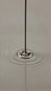Cinematic slow-motion macro video of a smooth, viscous beige-grey liquid pouring continuously into a calm surface. The thick silky cream creates mesmerizing soft concentric ripples and subtle ribbonin