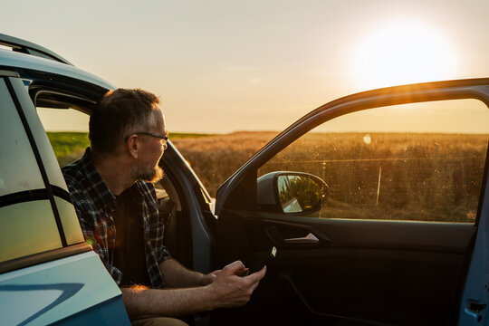 Middle-aged Caucasian farmer sitting in car with open door using smartphone at sunset. Smart farming concept.