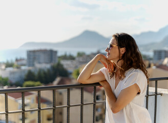 Obraz premium Beautiful young woman standing on apartment balcony and enjoying sunny summer day. City view background.