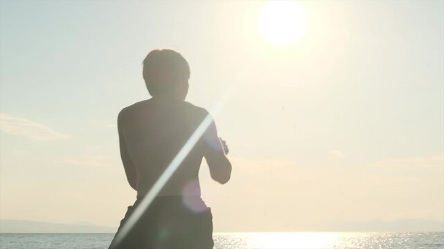 Young man trains MMA on the seashore at sunrise, performing a flying knee in slow motion during shadow boxing. Muay Thai striking technique, outdoor martial arts training.