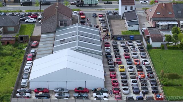 Slow drone footage showing parked roadsters lined beside a roadway near a commercial venue, The aerial perspective captures organized parking, surrounding buildings, and daytime urban transportation.