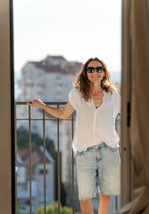 Obraz premium Beautiful millennial woman in sunglasses standing on balcony, looking at camera and smiling. View from inside apartment.