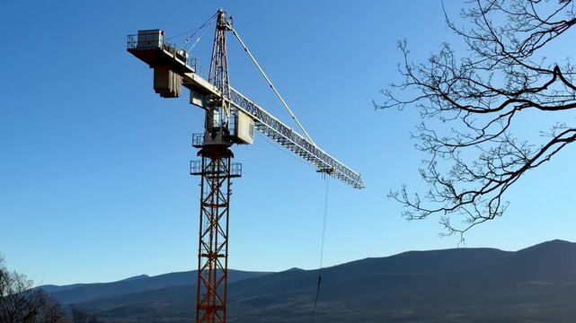A tall yellow construction crane rotating against a clear blue sky and distant mountain range, representing urban development, infrastructure projects, and industrial site progress