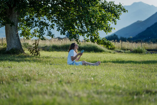 Young woman traveler using smartphone while sitting on green grass under large tree. Mobile connection in travel.