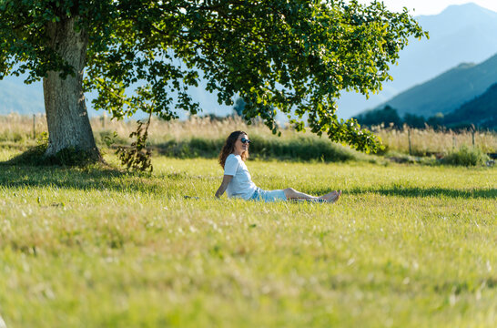 Beautiful young woman relaxing on green grass under a large tree, enjoying summer nature.