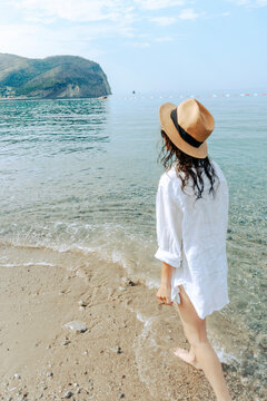Vertical back view of young woman in white shirt and hat walking on sandy sea beach.