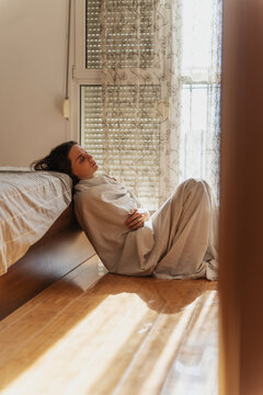 Sad lonely female feeling down, sitting on bedroom floor wrapped in bedsheet. Apathy, grief and despair.