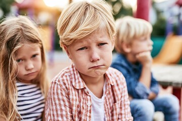 Fototapeta premium Three young children with sad expressions sitting outdoors, one boy looking directly at the camera with disappointment.