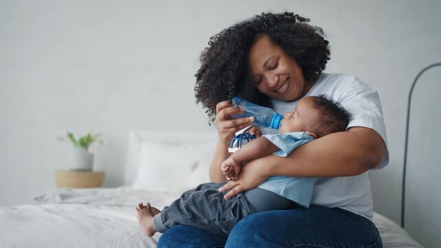 Cute healthy baby drinking water or formula from bottle, mother feeding child. Motherhood and childhood, african american mom practice bottle-feeding, enjoy happy family moments, calm scene at home