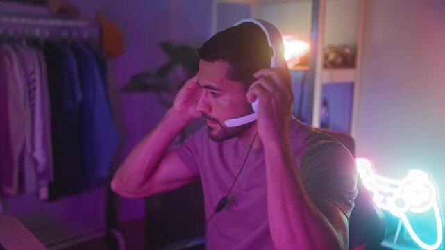 Man with headset and gamepad reaches to adjust microphone while smiling and gaming at a computer desk in a studio with neon controller sign; concentration.