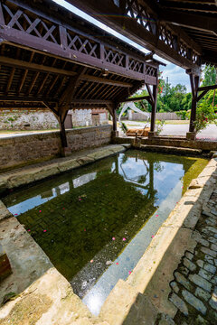 Interior view of a rustic wooden and stone wash-house with a reflective water basin scattered with rose petals, exposed timber roof framing, and a glimpse of a wedding canopy and straw bale outside.