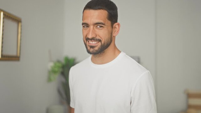 Man smiling with visible face and beard in a studio apartment interior wearing a white tshirt; friendly confidence.