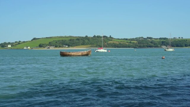 A simple small wooden fishing boat gently sways on the waves on a sunny day in a bay near big city, other yachts and houses on the shore are visible in the distance, video of a boat at sea