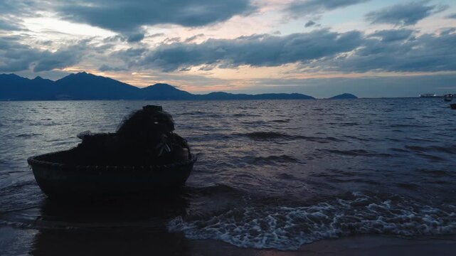 Traditional vietnamese coracle boat or basket boat resting on the shore near da nang at sunset, mountains and clouds creating a dramatic coastline silhouette against the tranquil ocean