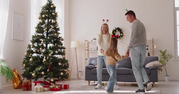 Christmas family dancing fun dance in circle holding hands at home by Xmas tree with gift boxes. Parents spin with child, share laughter and festive mood, winter holiday celebration in living room