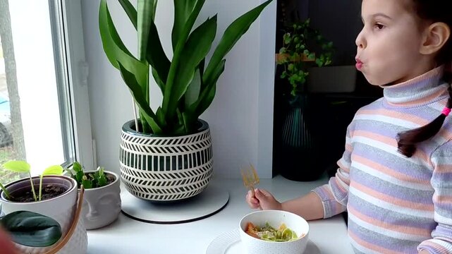 A little girl eats pasta with vegetables near the window. Funny child eats vegetables and laughs