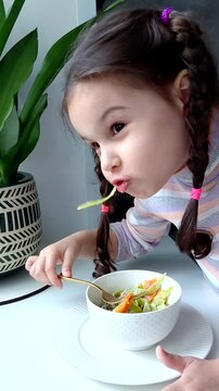 A little girl eats pasta with vegetables near the window. Funny child eats vegetables and laughs