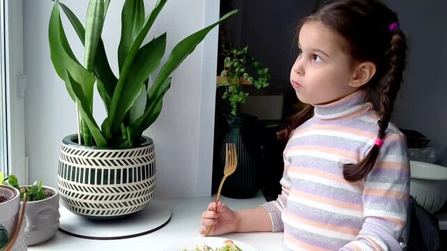 A little girl eats pasta with vegetables near the window. Funny child eats vegetables and laughs