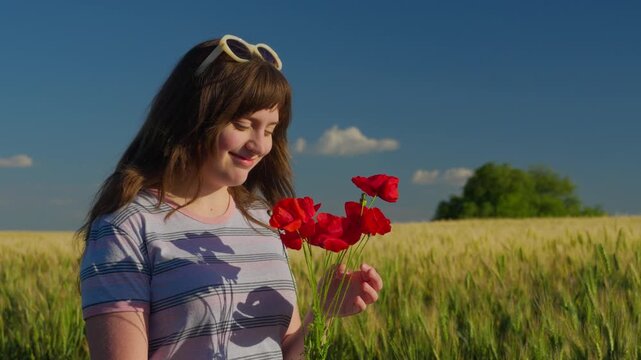 A smiling teenage girl enjoying nature while holding bright red poppy flowers in a golden grain field under a clear blue sky at sunset