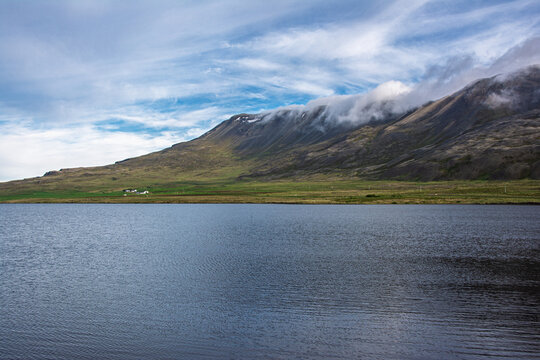 Icelandic landscape presenting a calm lake reflecting the sky, with cloud formations clinging to the slopes of green and brown mountains, evoking a sense of tranquil nature and travel